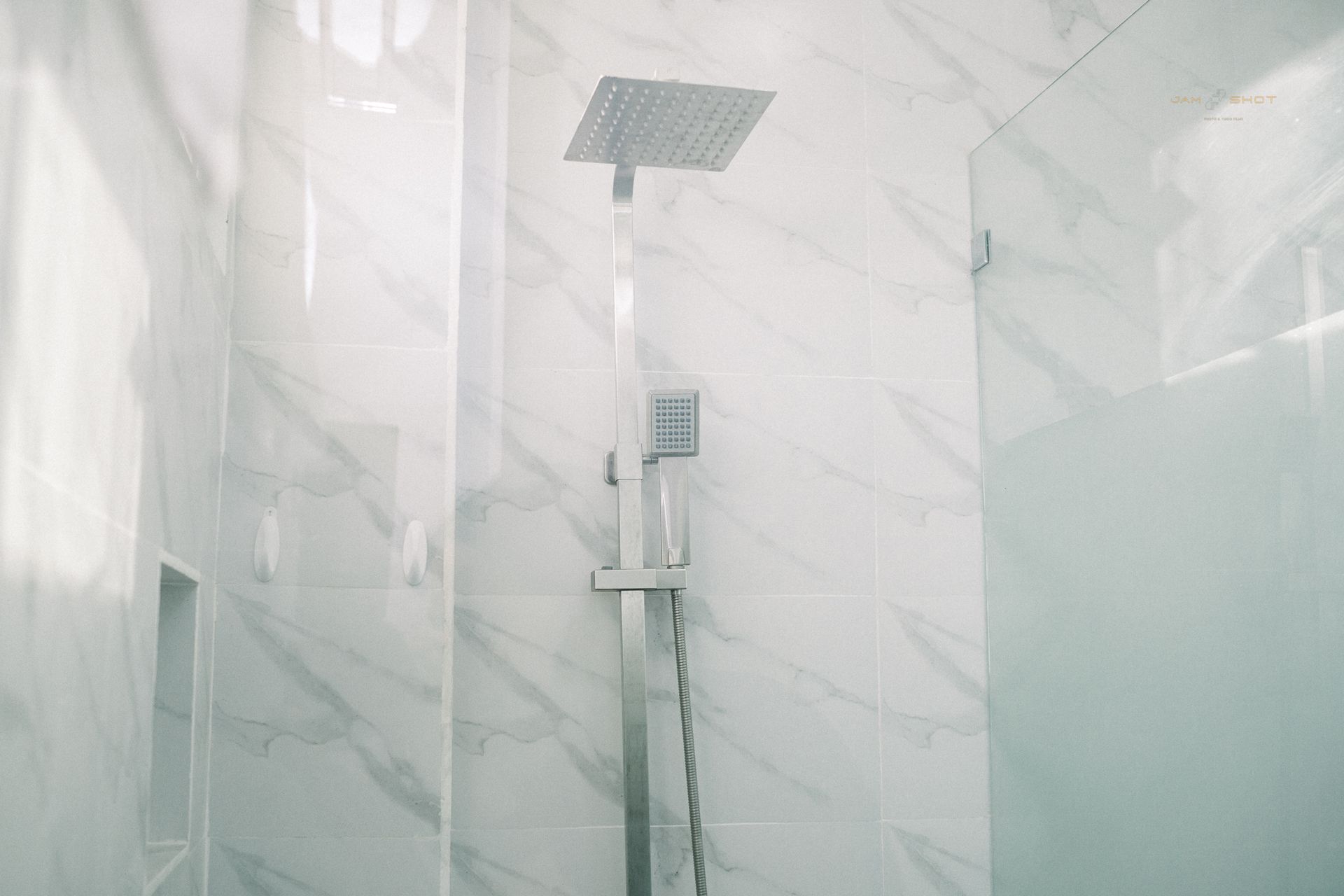 A modern, square rainfall showerhead and wall-mounted metal fixtures against white marble-patterned tiled walls.