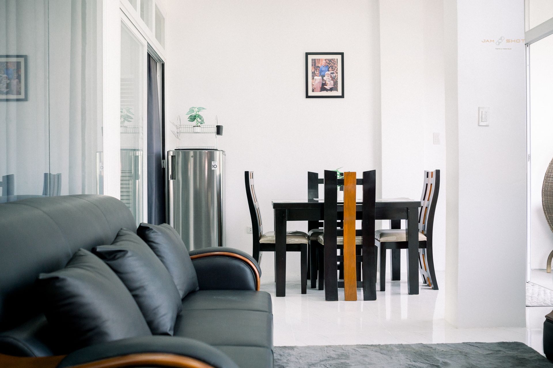A modern interior showing a dark grey sofa in the foreground, with a dining table set and a refrigerator in the back.