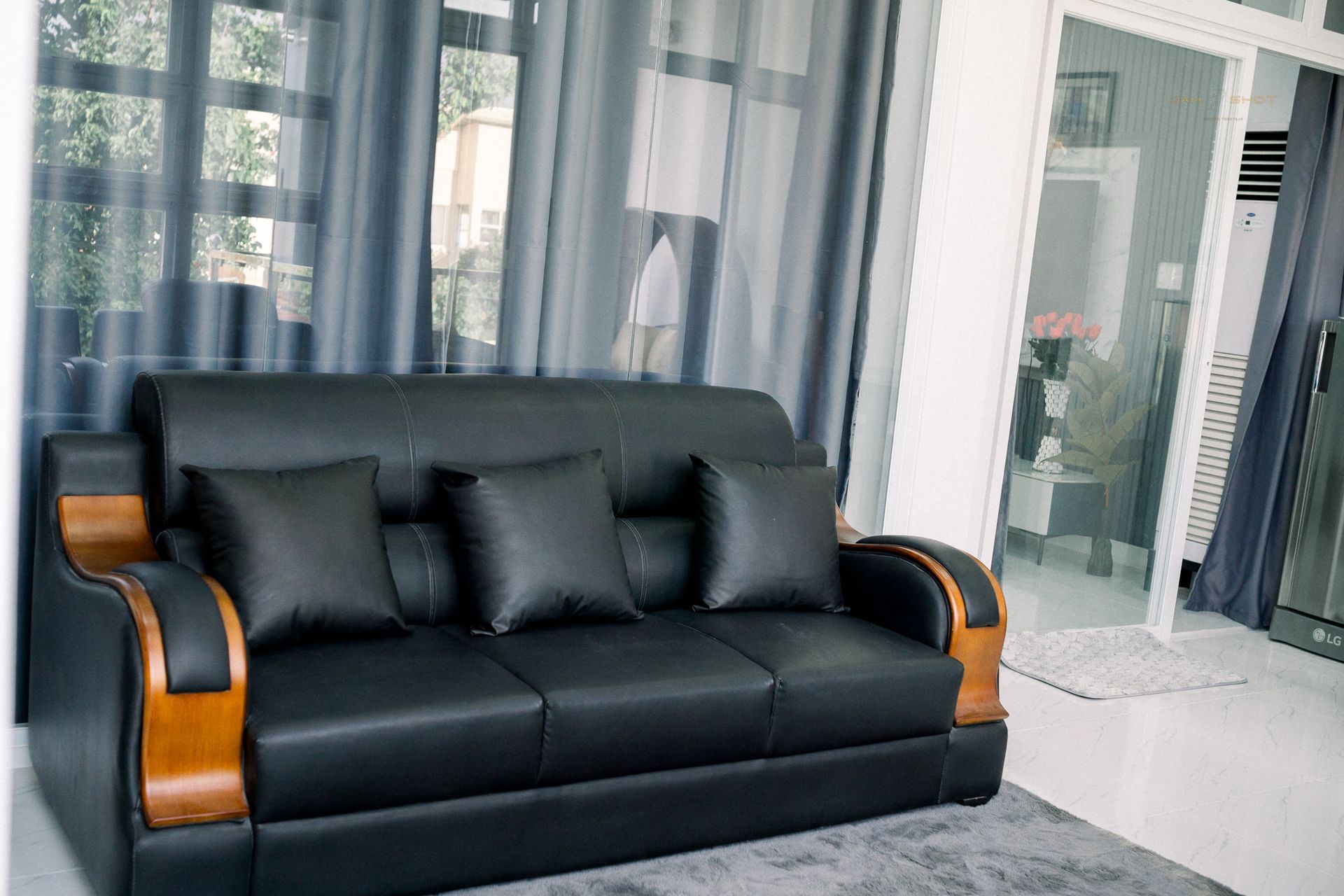 A black, three-seater leather sofa with wooden armrests, featuring three throw pillows, set against sheer grey curtains.