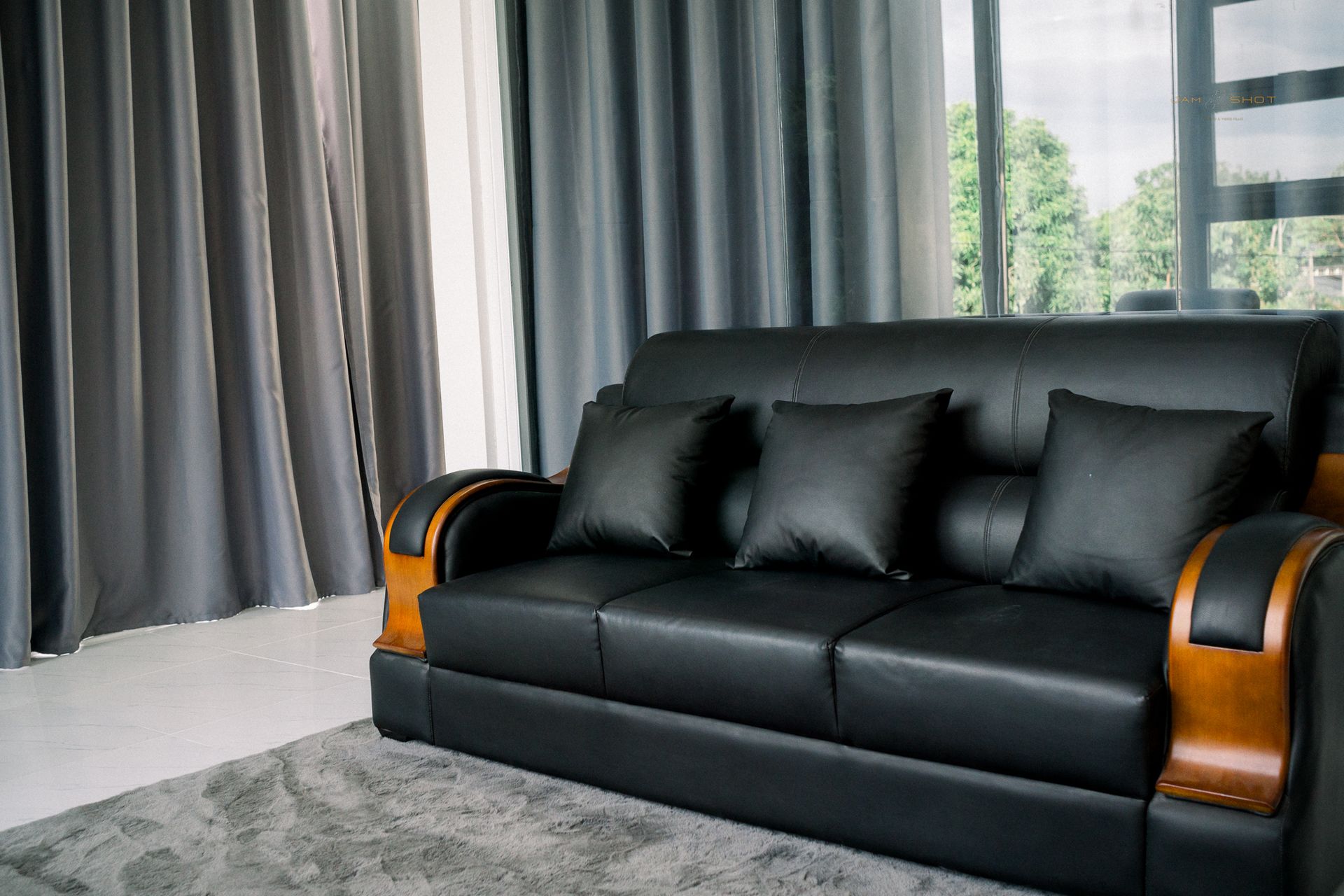 A black leather sofa with wooden armrests and three black throw pillows sits on a gray rug against gray curtains.