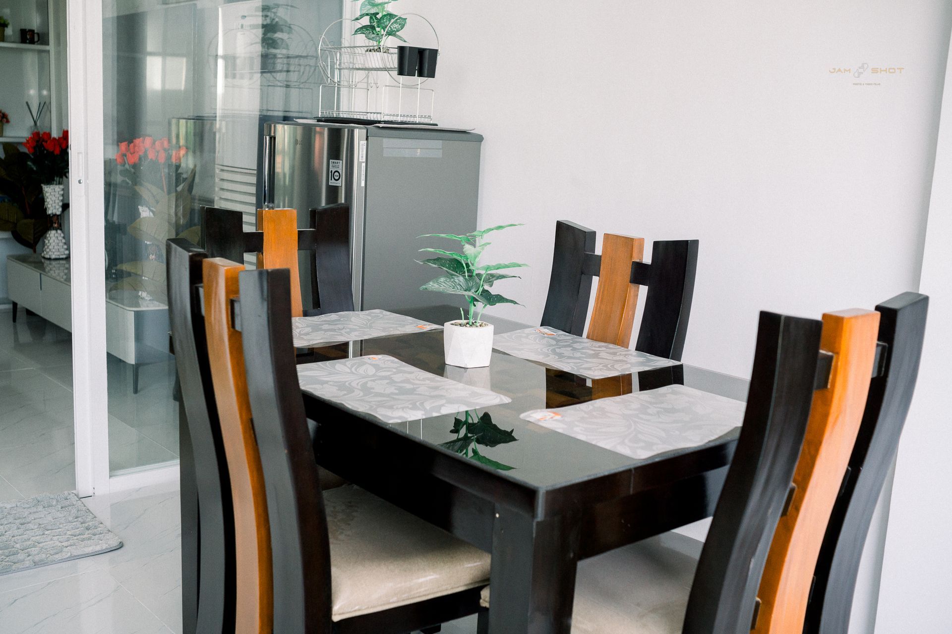 A modern dining set with a glass-topped table and four high-backed chairs in a white-walled room with a small plant.