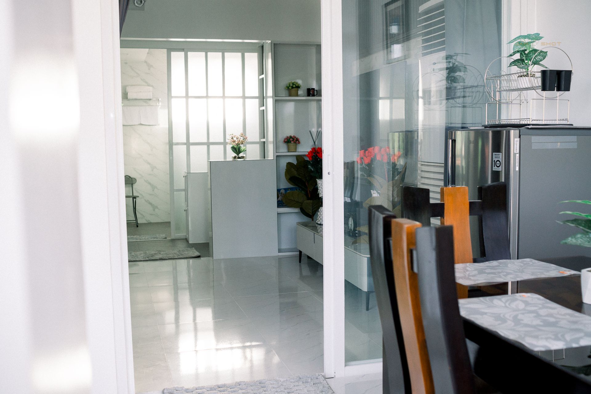 A dining area with a glass-topped table and chairs, overlooking a bright, minimalist room with shelving and tile floors.