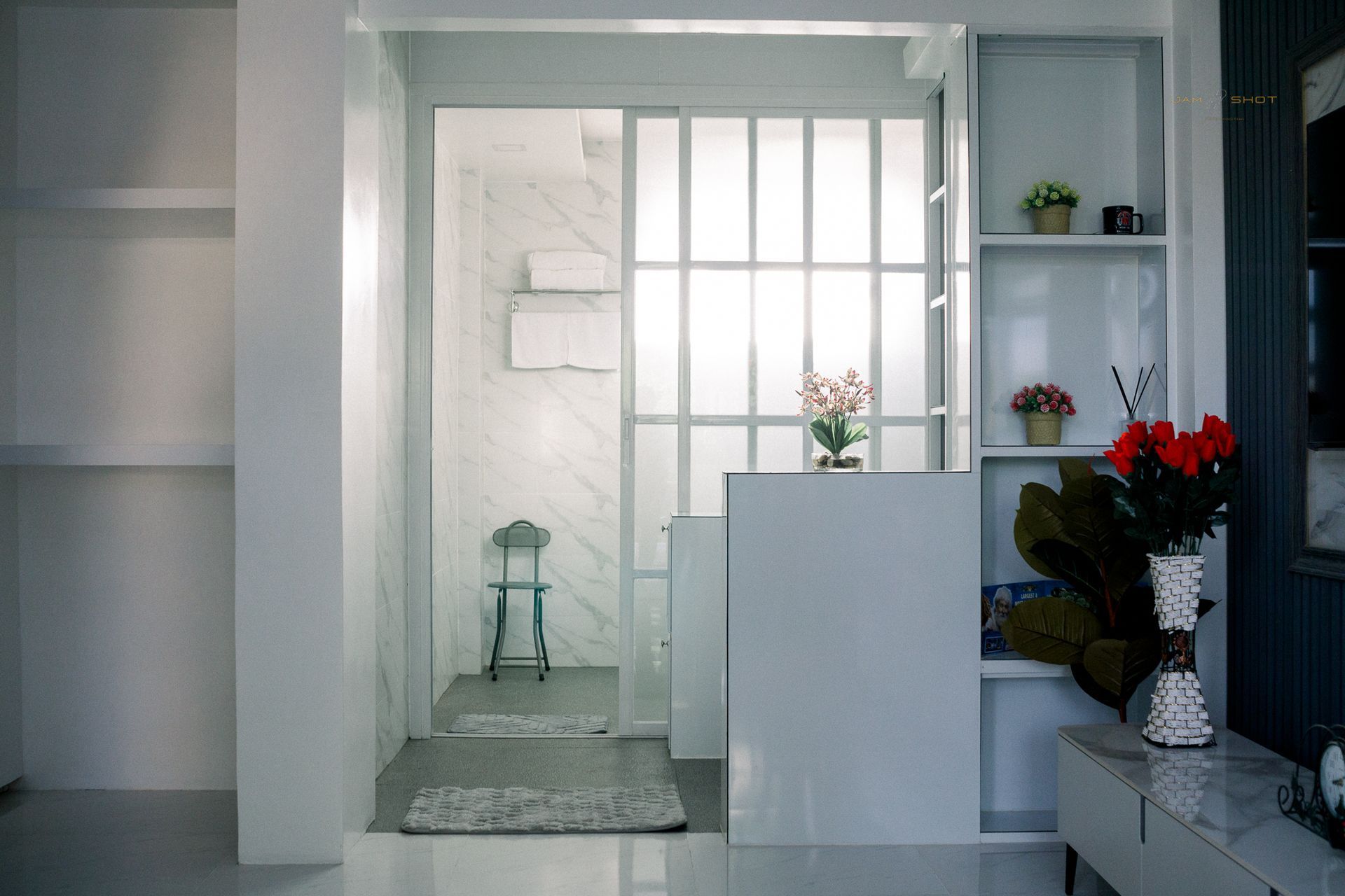 A bright, modern interior features a white glass partition leading to a small, marble-tiled room with a stool and towel rack.