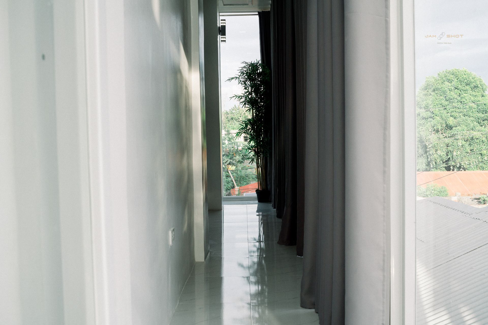 A sunlit hallway with white walls and a reflective tile floor, leading toward a glass door opening onto a leafy green view.