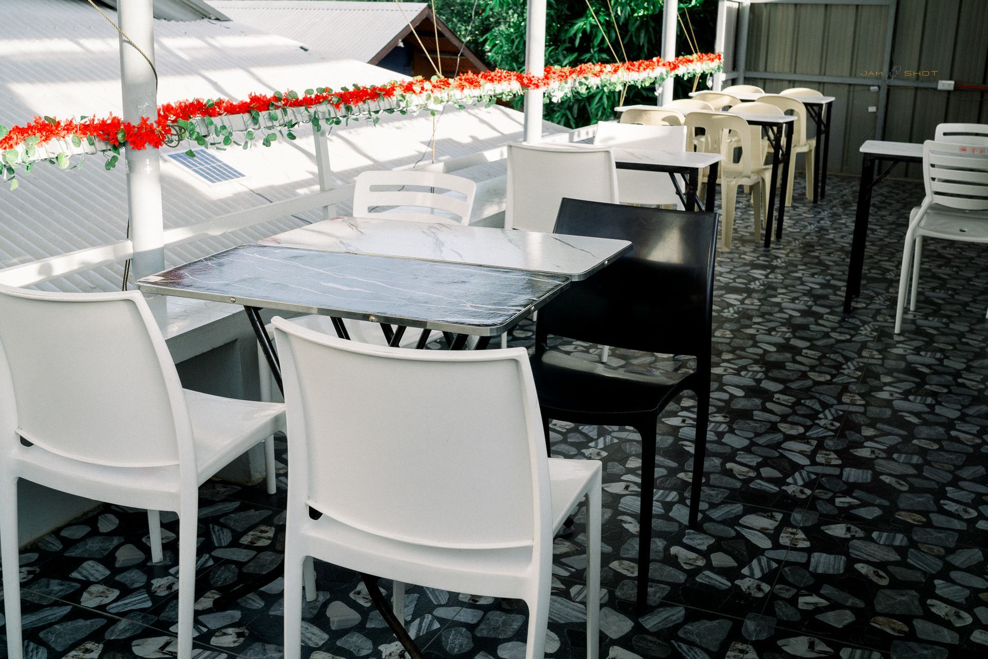 An outdoor patio dining area with white tables, chairs, and red flower garlands hanging from the roof.