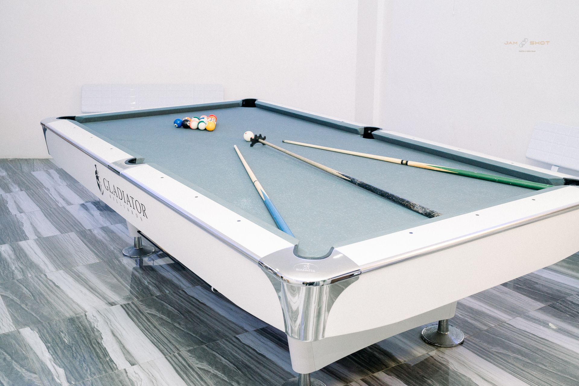 A white pool table with a grey felt surface, three cues, and a small cluster of balls, set on a grey patterned floor.