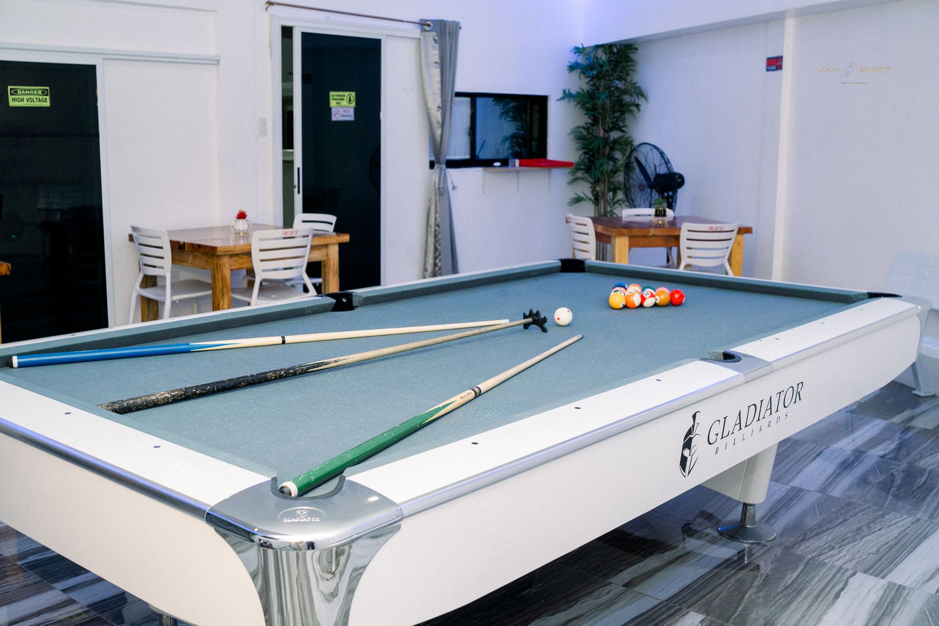 A white pool table with cue sticks and balls sits in a modern game room with white furniture and a potted plant.