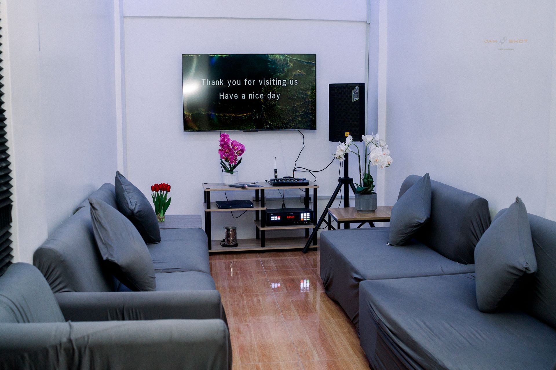 A cozy room with gray sofas facing a wall-mounted TV, a media console, a speaker on a tripod, and decorative flowers.