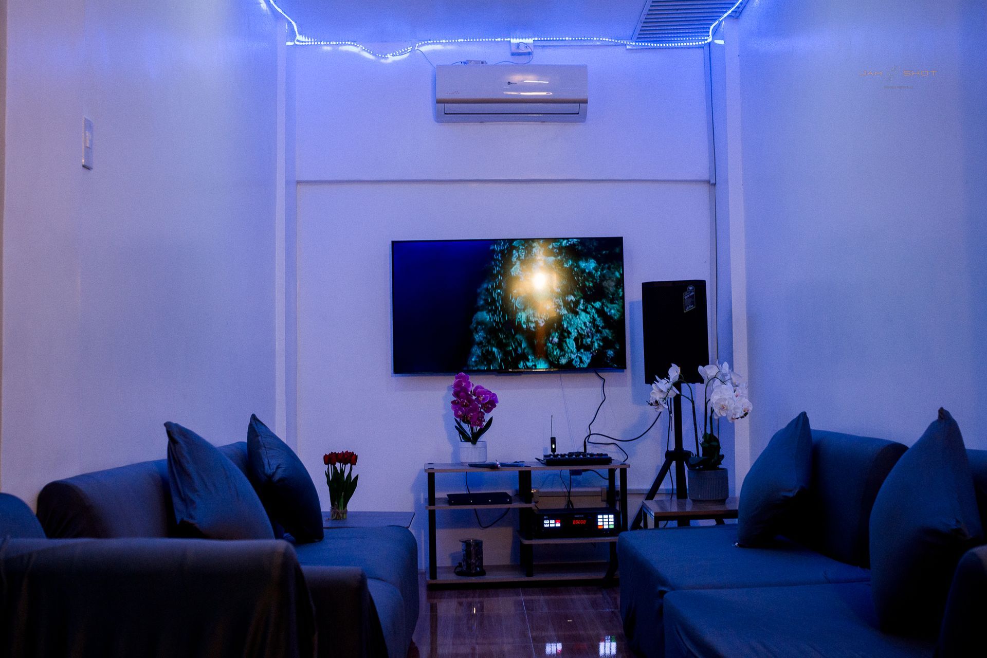A living room bathed in blue light, featuring two dark sofas, a wall-mounted television, and an air conditioning unit.