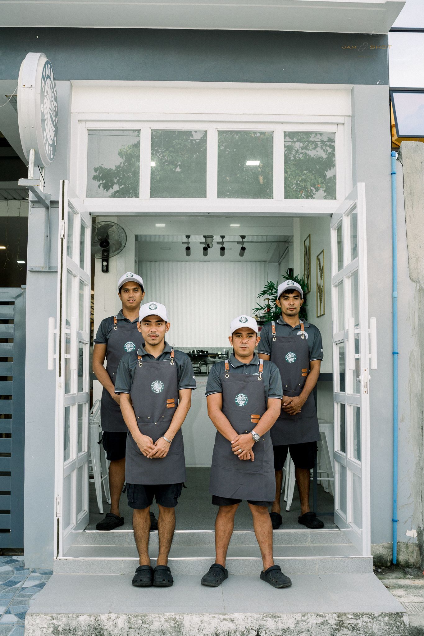 Four individuals in grey aprons and caps stand in a line at the entrance of a modern, grey-toned storefront.