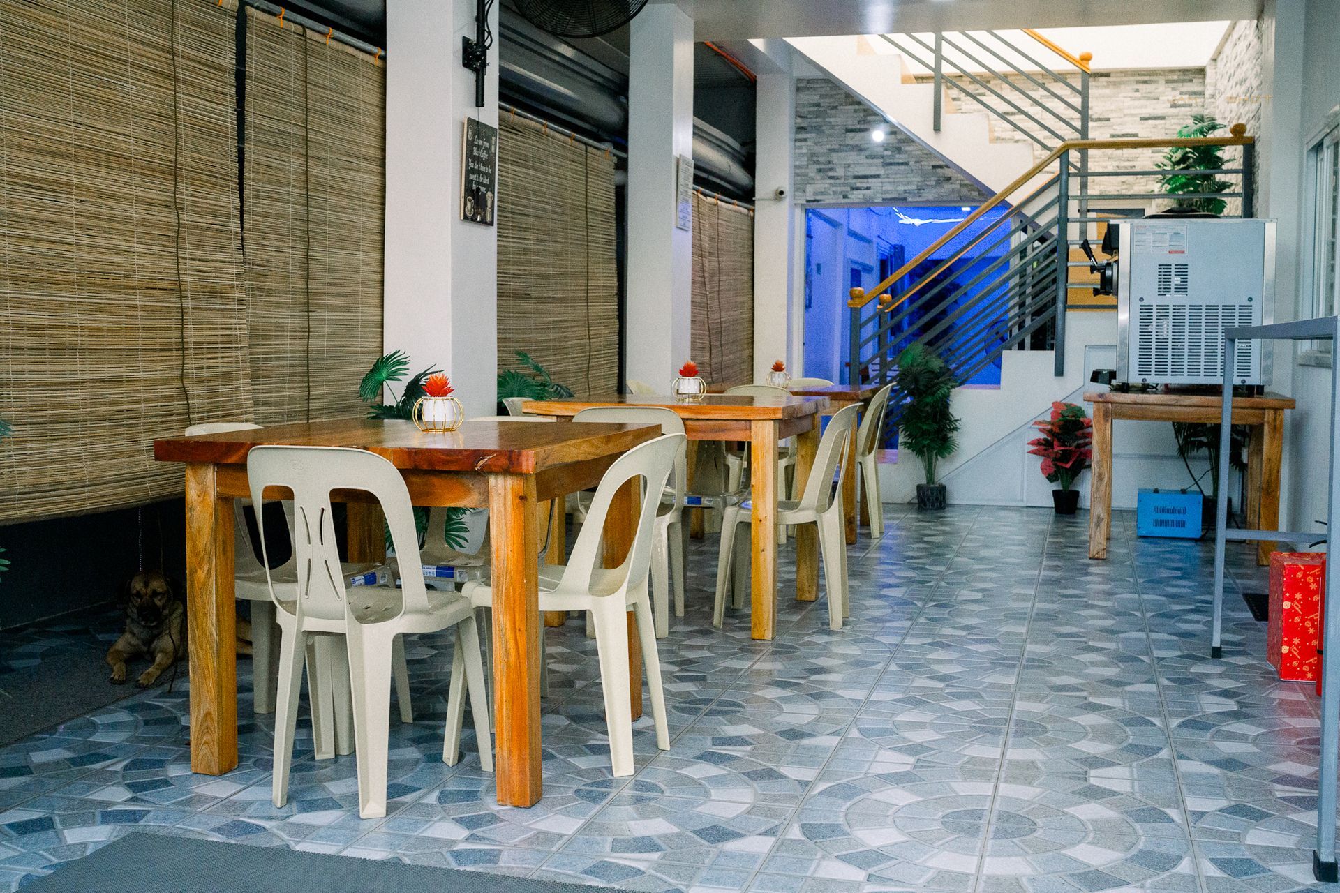 An interior view of a dining area with several light-colored plastic chairs arranged around wooden tables on a tiled floor.