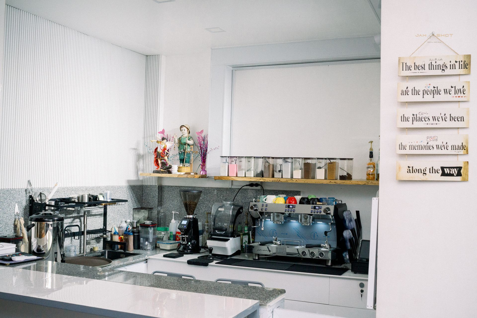 A kitchen counter area with a coffee machine, grinder, and ingredients in clear containers, featuring wall-mounted signs.