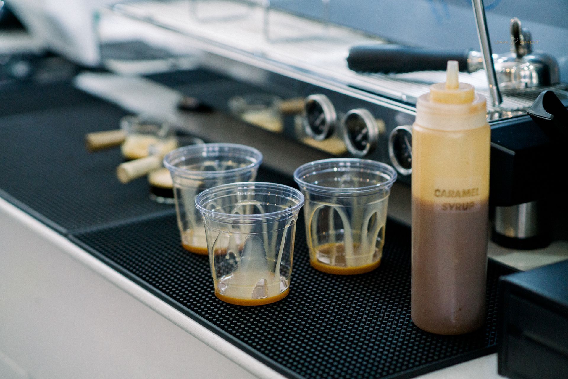 Three clear plastic cups with caramel drizzle inside sit on a black service mat next to a bottle of caramel sauce.