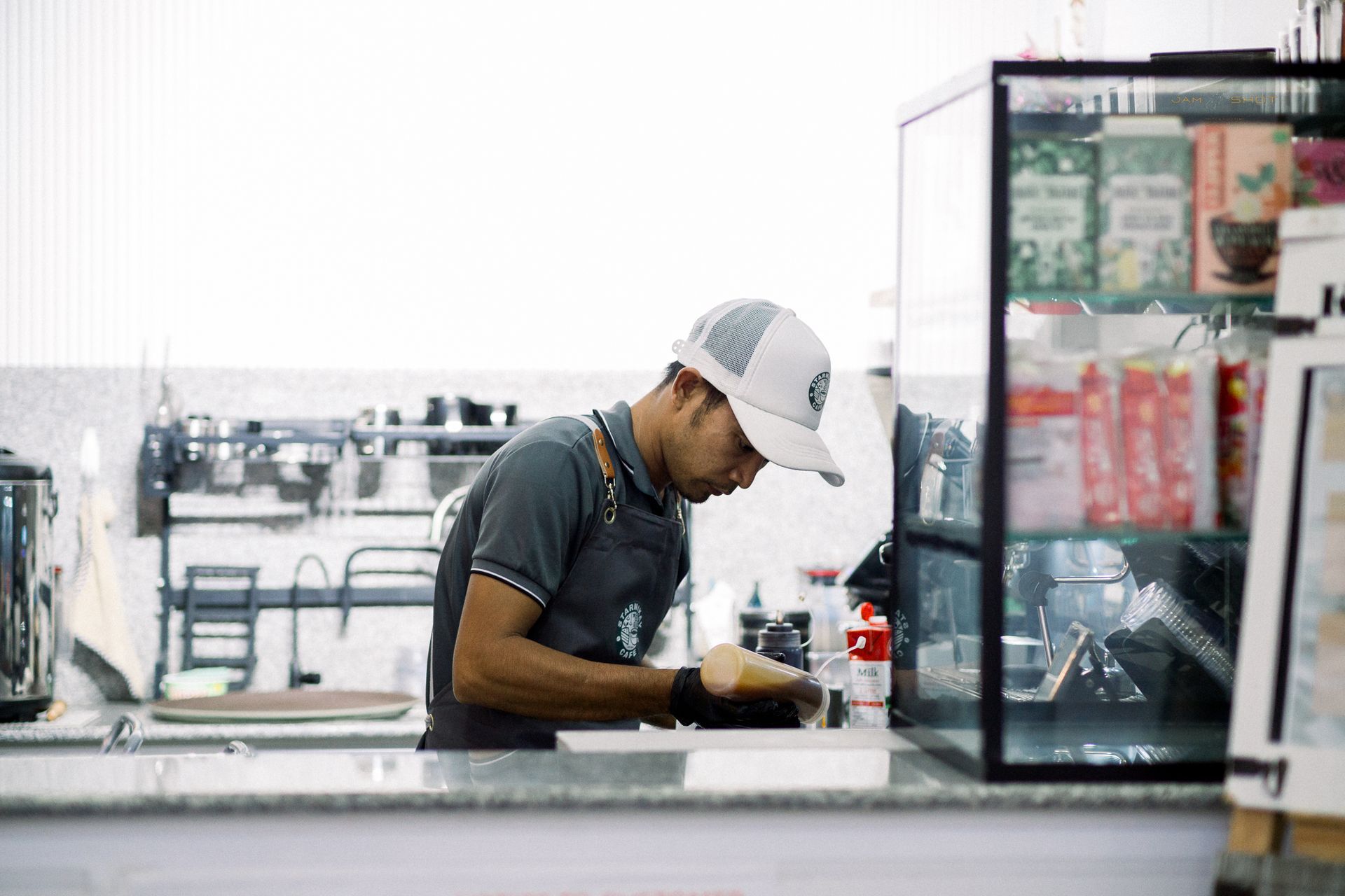 A barista wearing a cap and apron prepares food or drink behind a coffee shop counter with display cases.
