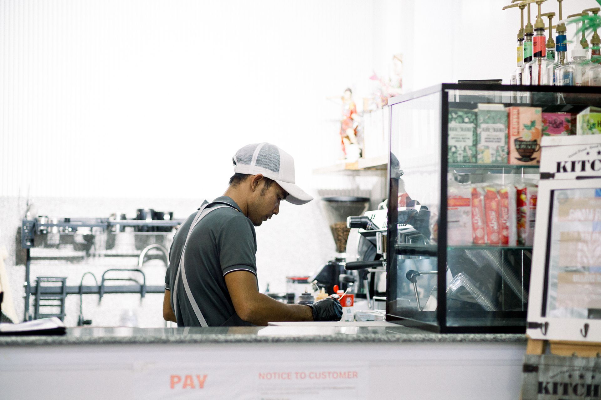 A person in a gray shirt and cap wearing black gloves works behind a cafe counter next to a glass display case.