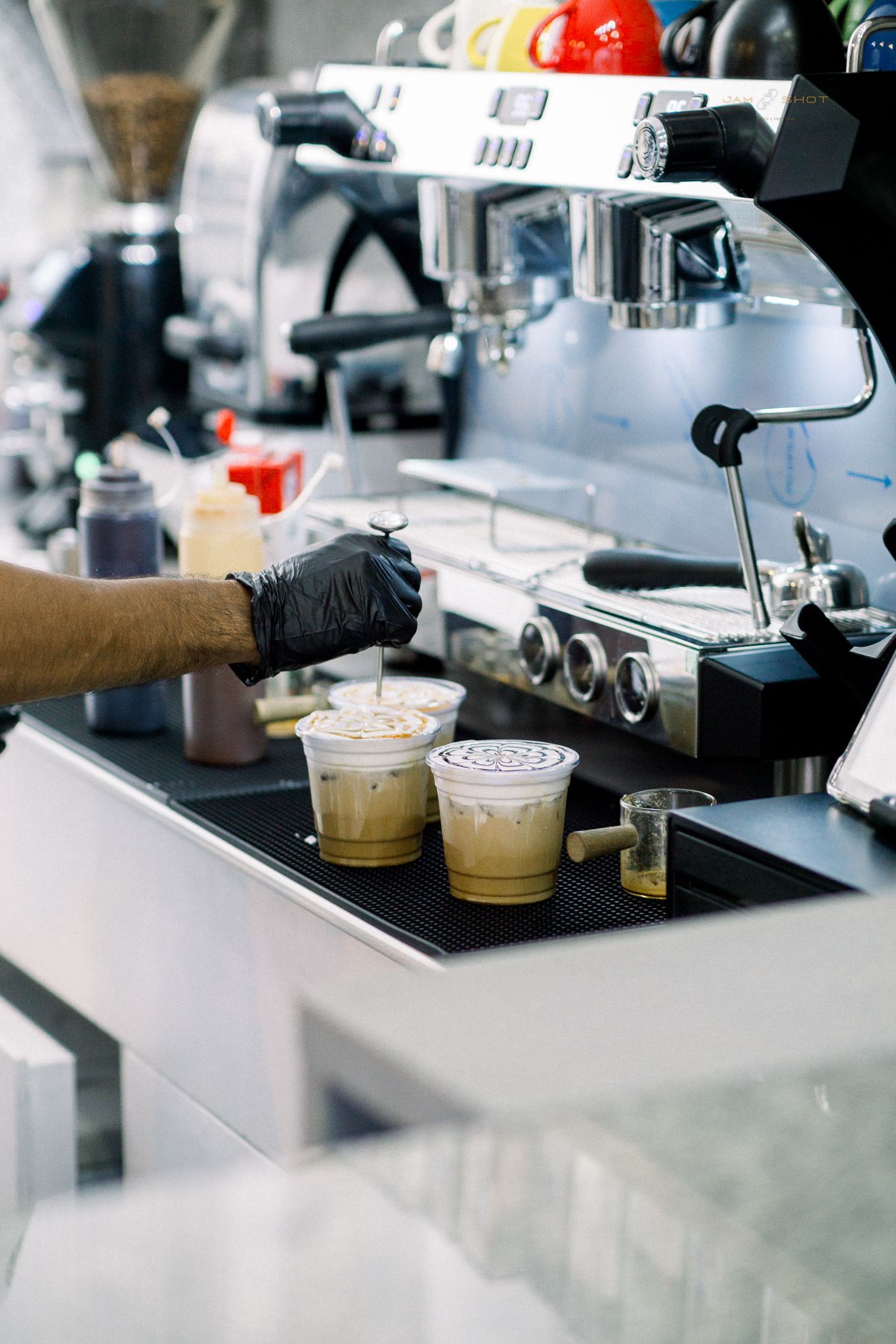 A barista wearing a black glove uses a small tool to craft toppings on two iced coffee drinks by an espresso machine.
