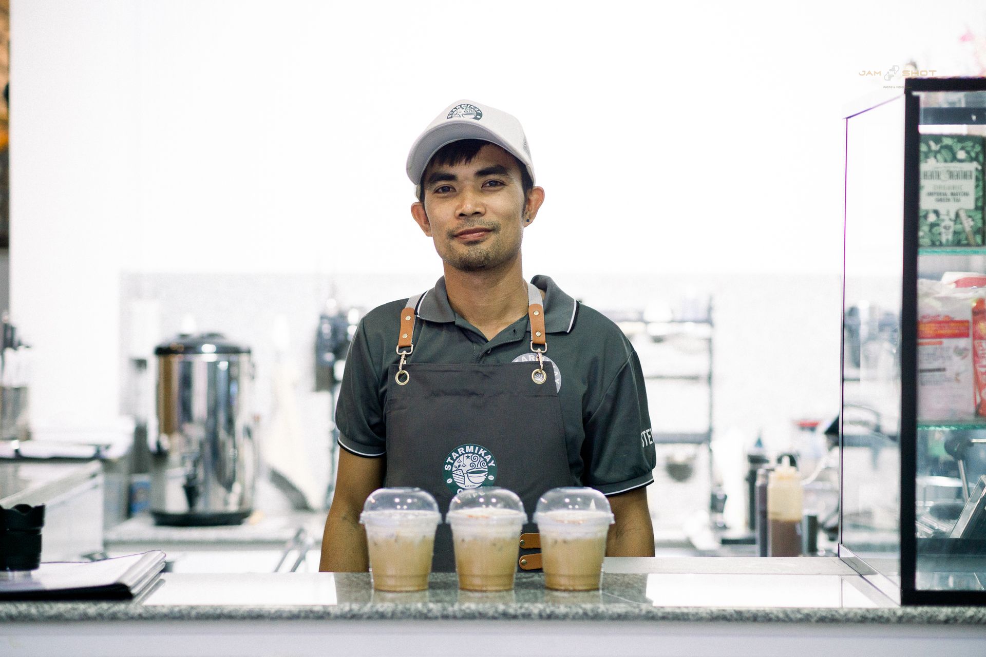 A barista in a gray uniform and cap stands behind a counter with three iced coffee drinks in plastic cups.