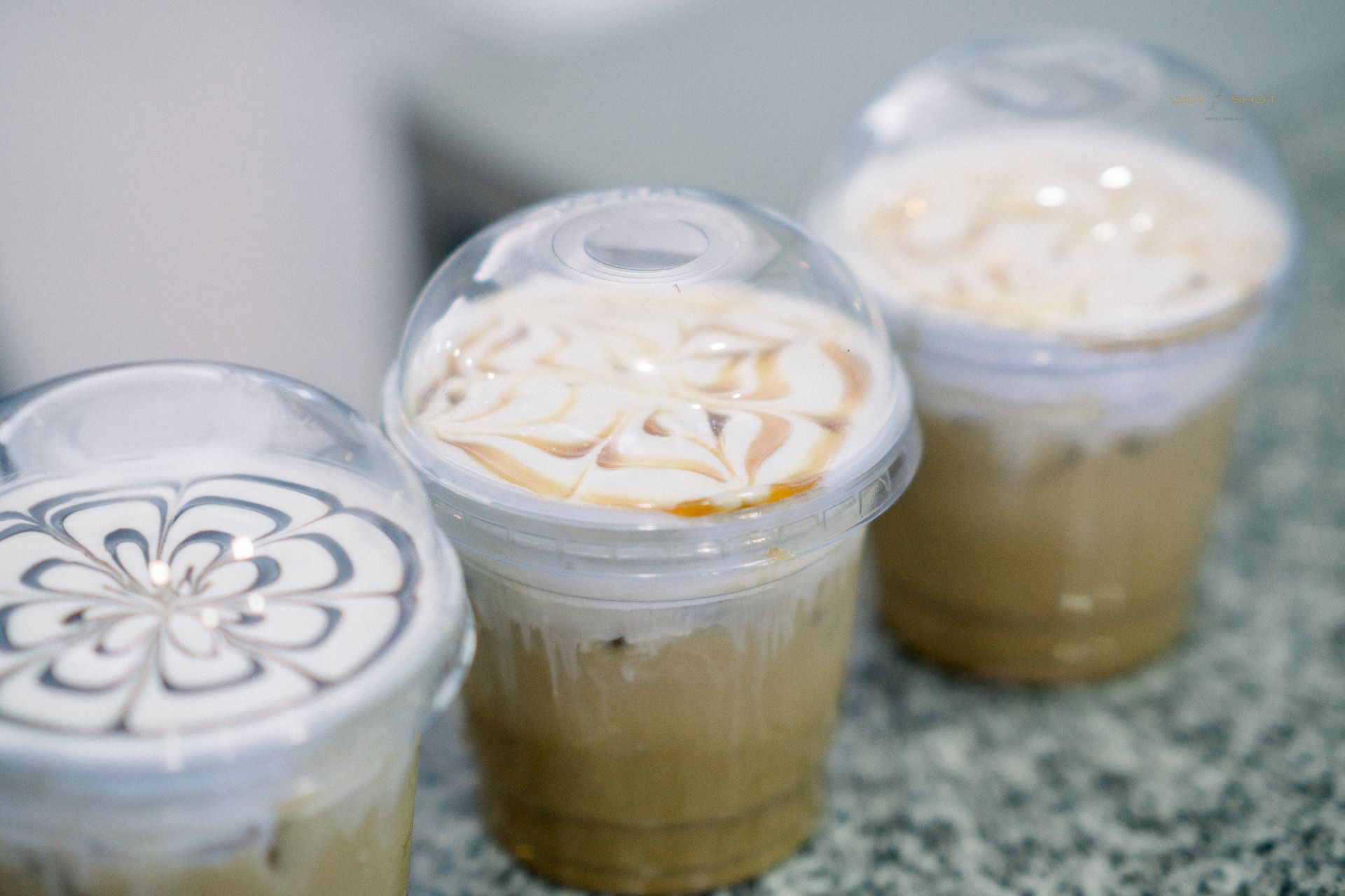 Three clear plastic cups of iced coffee with decorative foam art on the lids, sitting on a speckled countertop.
