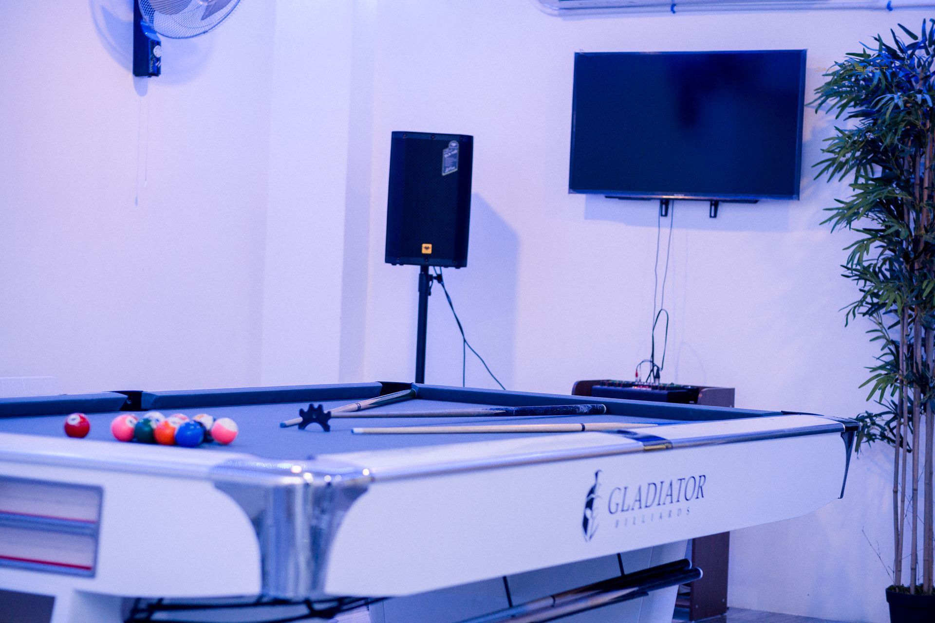 A white pool table with colorful balls and cues, positioned under a speaker and television in a bright, modern room.