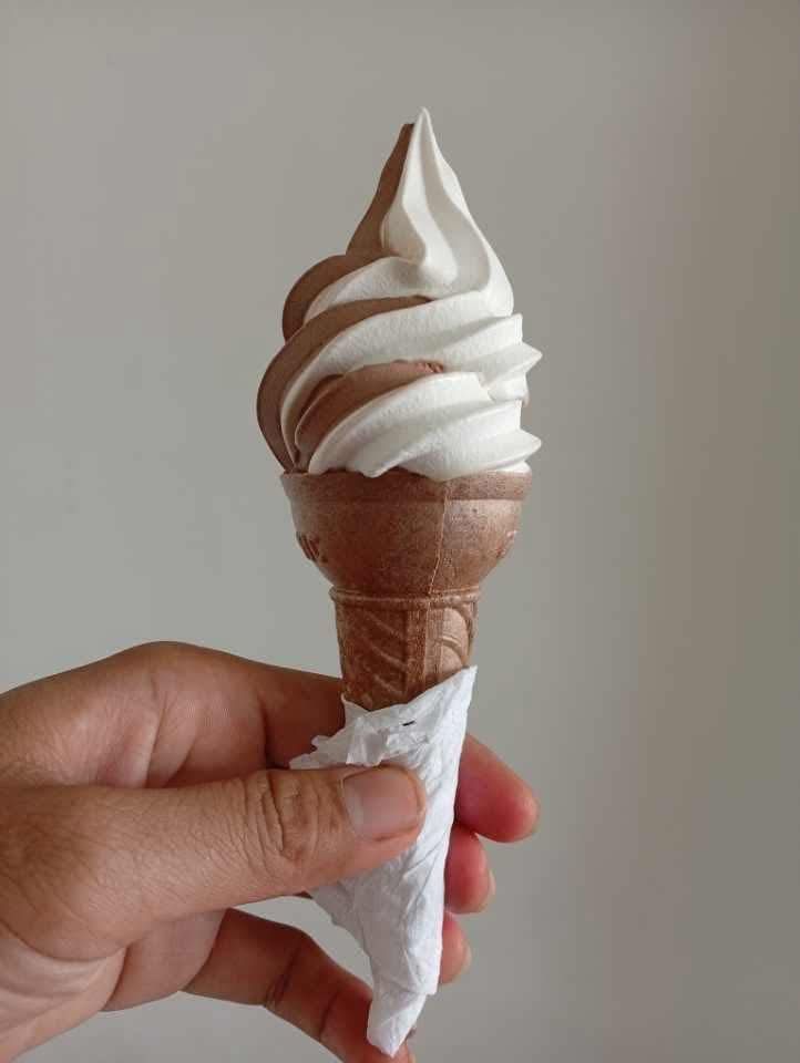 A hand holds a chocolate and vanilla swirl soft-serve ice cream cone wrapped in a paper napkin against a plain wall.