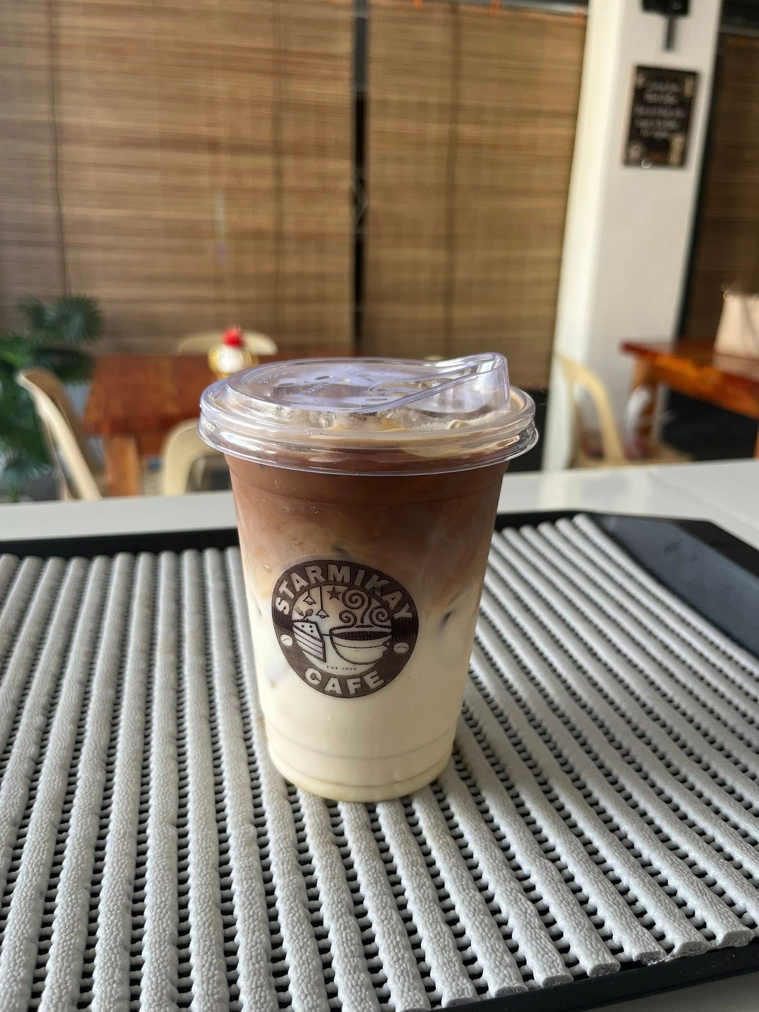 A plastic cup of iced coffee with a layered white and brown appearance, sitting on a gray mat in a cafe setting.