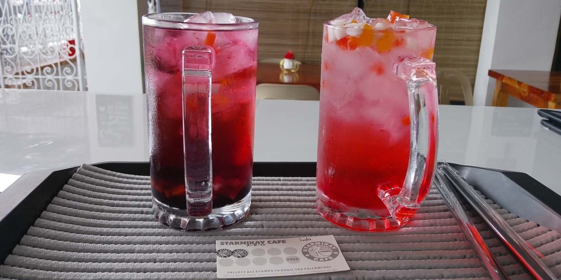 Two glass mugs filled with ice and vibrant red drinks sit on a textured gray placemat with a business card and straws.