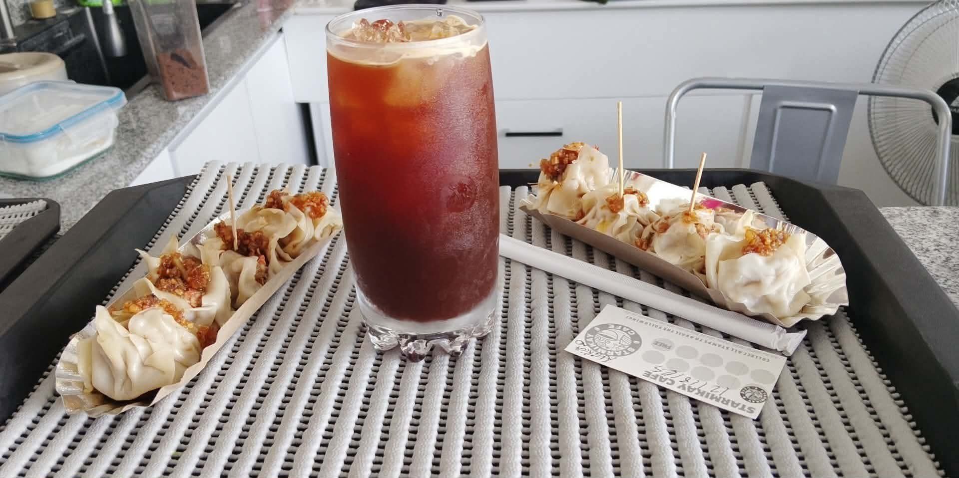 Two rows of siomai with fried garlic garnish on a ridged tray, alongside a tall glass of iced dark coffee on a counter.