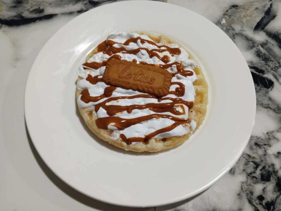 A circular waffle topped with whipped cream, drizzled with caramel sauce, and finished with a Lotus Biscoff cookie.