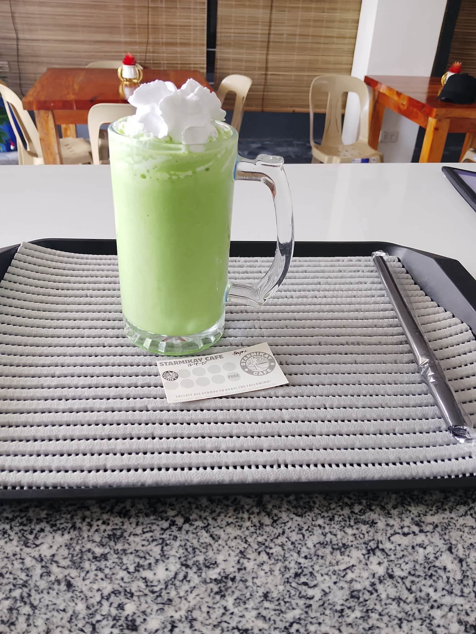 A glass mug of light green milkshake topped with whipped cream on a tray, set on a counter in a café.