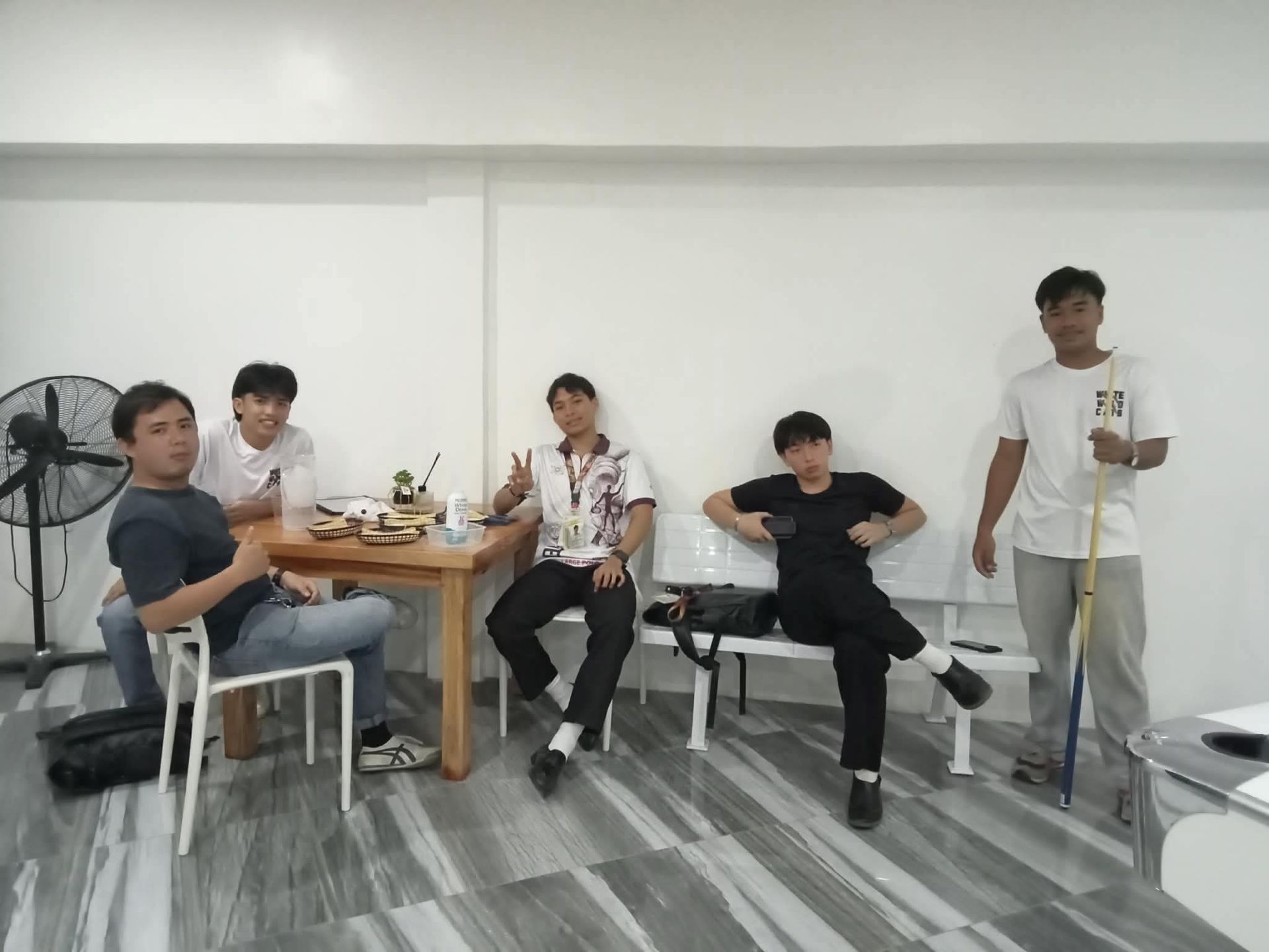 Five people gather in a brightly lit room with white walls, four seated at a wooden table and one standing with a cue.