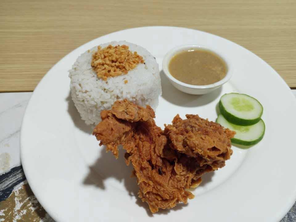 A plate with fried chicken, rice topped with crispy garlic, a side of gravy, and two cucumber slices.