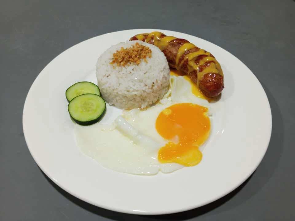 A plate with a scoop of rice topped with crispy garlic, a fried egg, a drizzled sausage, and two cucumber slices.