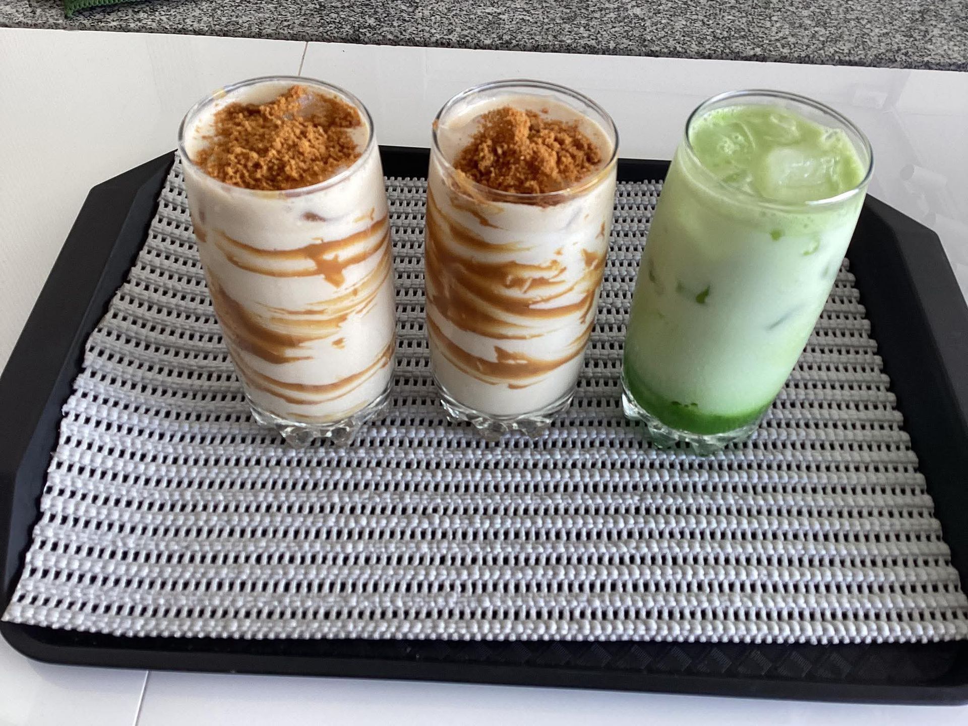 Three glasses on a black tray: two with white, swirled cream topped with brown crumbs, and one green iced beverage.