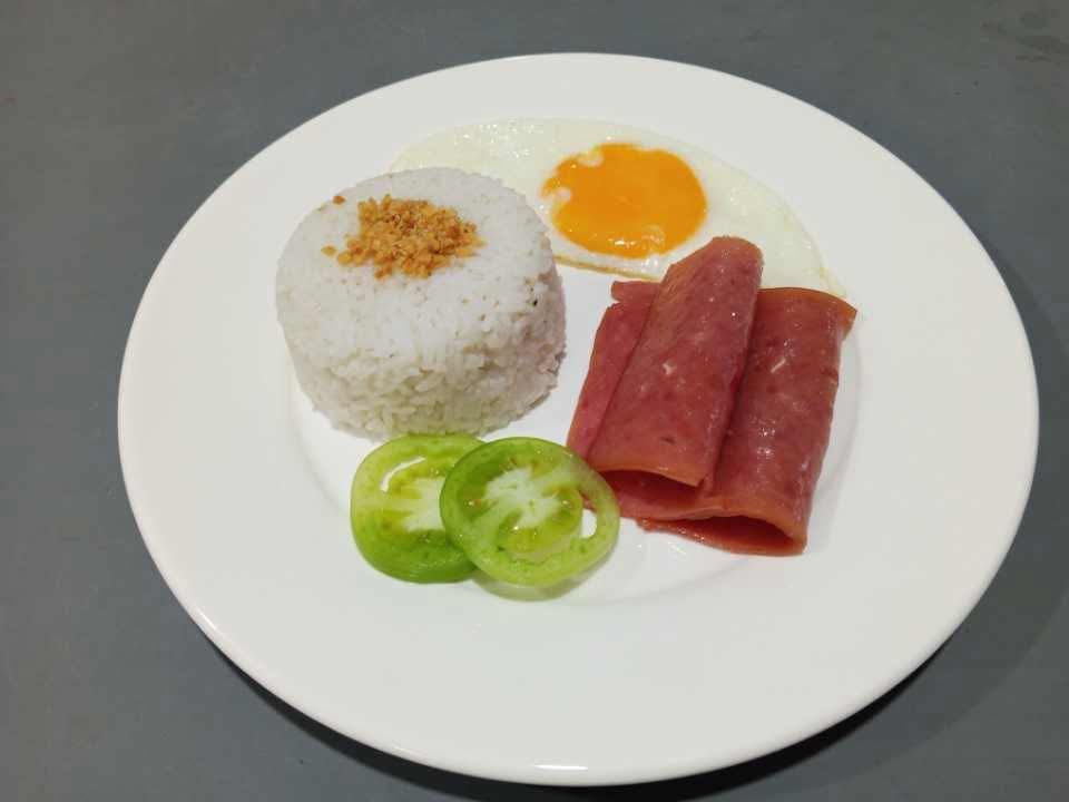A white plate with a scoop of garlic rice, a fried egg, slices of luncheon meat, and two slices of green tomato.