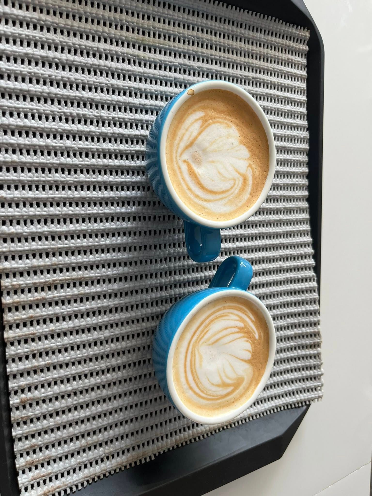 Two blue mugs of latte art coffee sit on a gray textured mat placed on a dark tray.