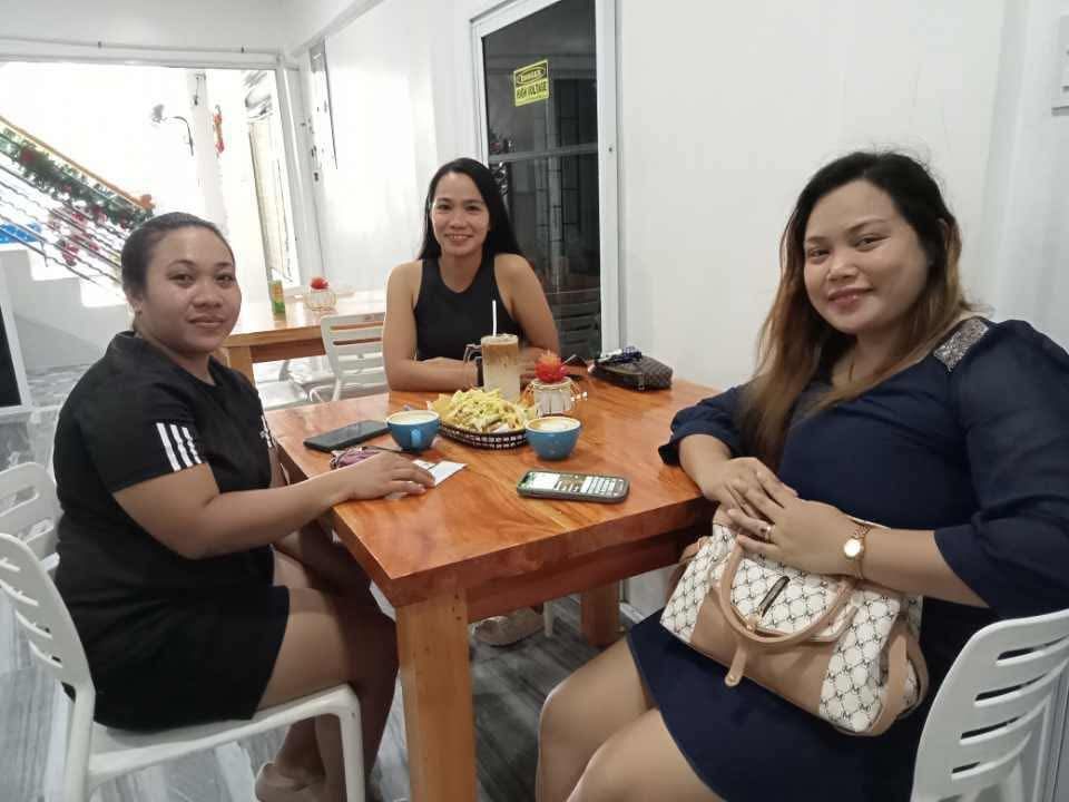 Three people sit at a wooden table in a bright room, posing for a photo with a shared snack and drinks.