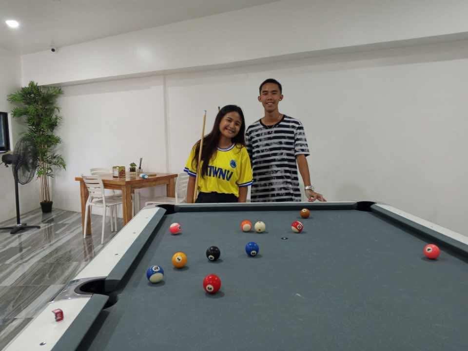 Two people stand behind a pool table in a bright room, one wearing a yellow shirt and the other a striped shirt.