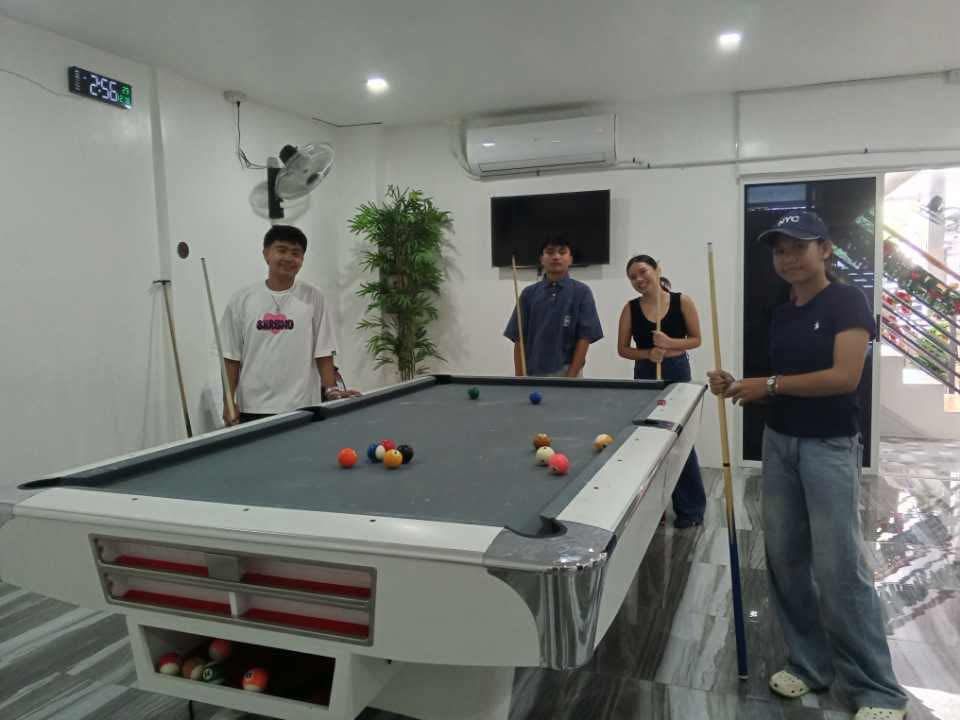 Four people pose around a billiards table in a bright, modern room, some holding cues and preparing to play.