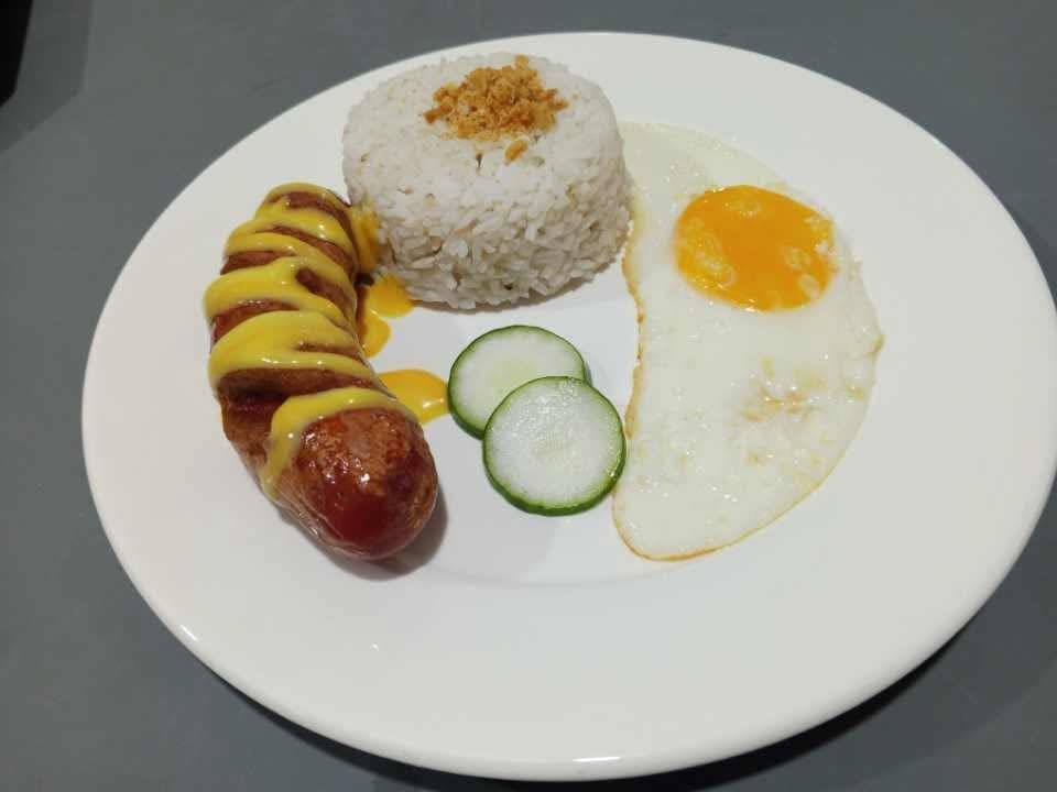 A plate with a sliced sausage topped with mustard, a scoop of garlic rice, a fried egg, and two cucumber slices.