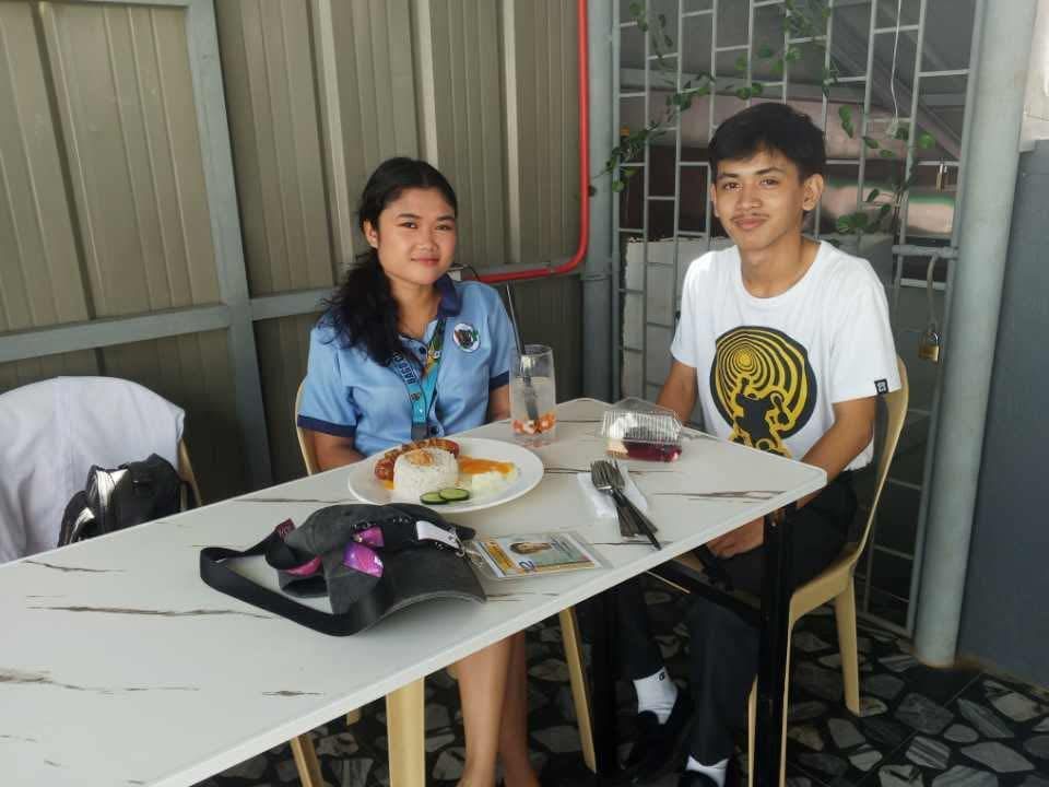 Two people sit across from each other at a table with plates of food, smiling at the camera in a covered outdoor area.