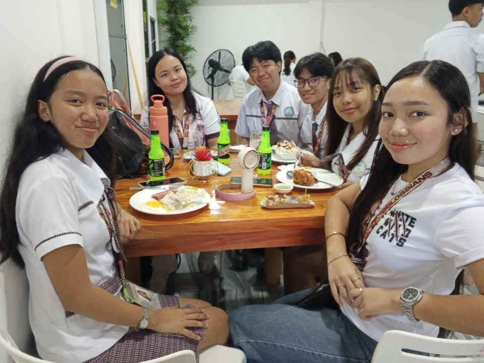 A group of students in uniforms sit around a wooden table, smiling while eating together in a bright, casual setting.