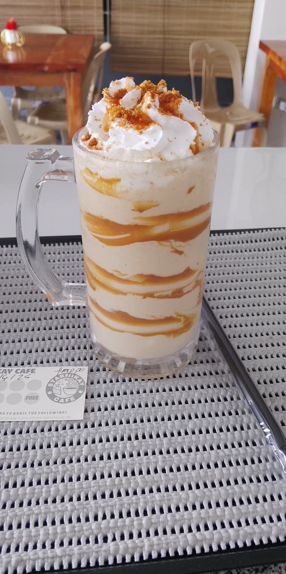 A tall glass mug filled with a creamy, caramel-swirled blended drink, topped with whipped cream and caramel drizzle.