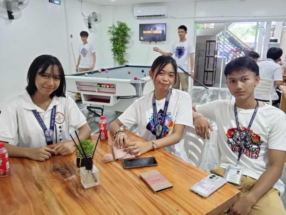 Three people in white shirts sit at a wooden table in a room with a pool table and a television in the background.