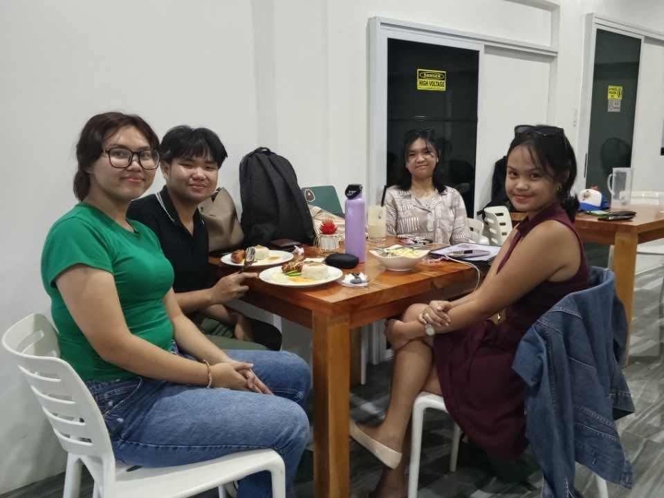Four people sit at a wooden table in a bright room, eating a meal together.