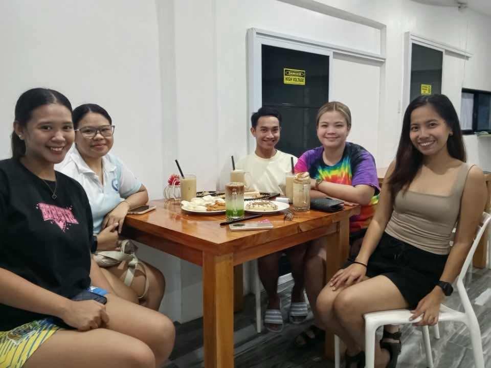 Five people smile while seated around a wooden table in a brightly lit indoor space with drinks and plates of food.