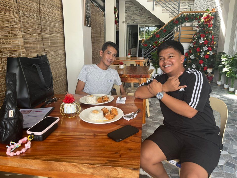 Two people sit at a wooden table in an outdoor dining area, smiling and posing with meals, near a decorated Christmas tree.
