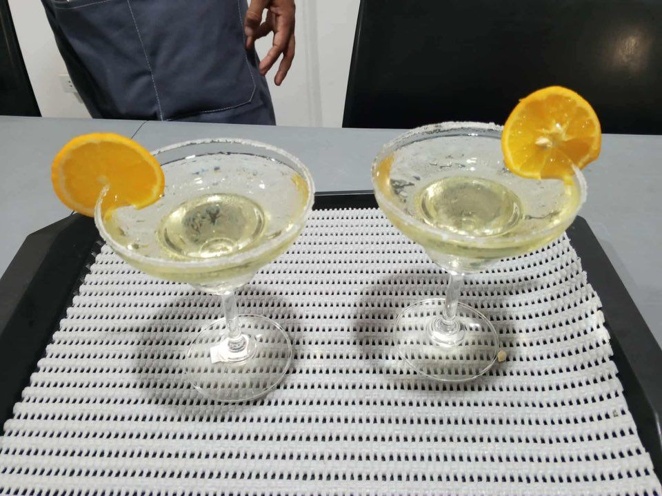 Two margaritas in stemmed glasses garnished with orange slices sit on a white mat on a black tray.