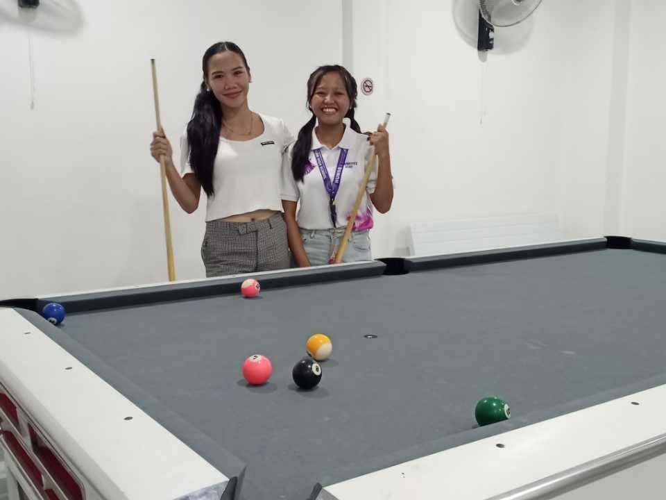 Two people stand smiling behind a pool table with several colored balls scattered on the gray felt.