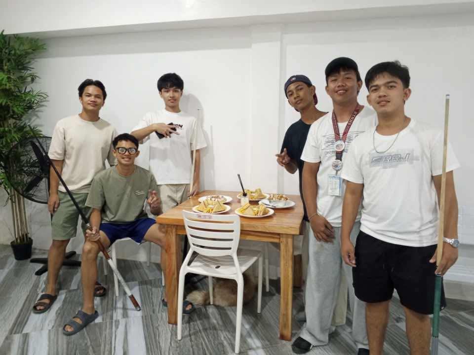 A group of seven people pose indoors near a table with food, with one person holding a pool cue and another a fan.