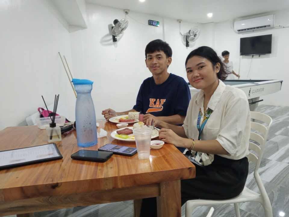 A person and a peer sit at a wood table in an indoor lounge, eating together with a blue water bottle and phones nearby.